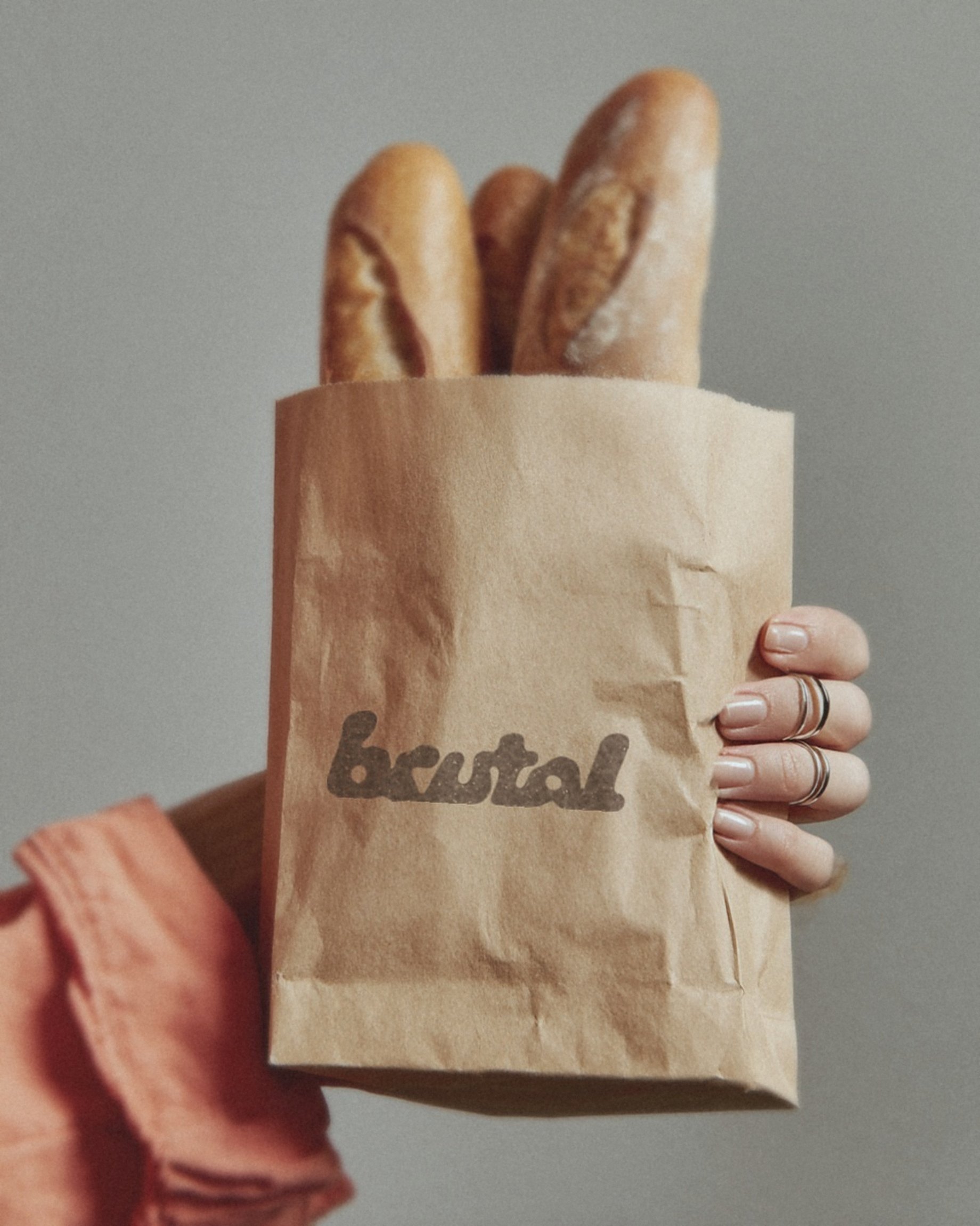 A hand with silver rings holding a kraft bread bag stamped with the BRUTAL logo in brown ink.