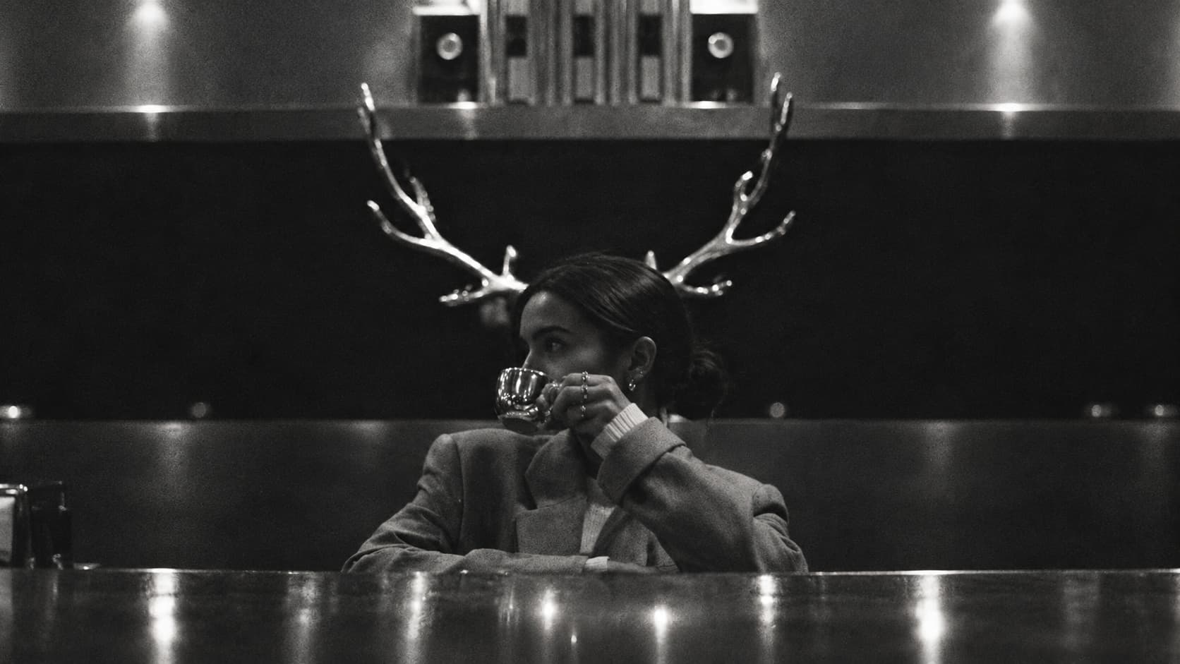 A woman at the bar drinking coffee, metal antlers on the wall behind her head