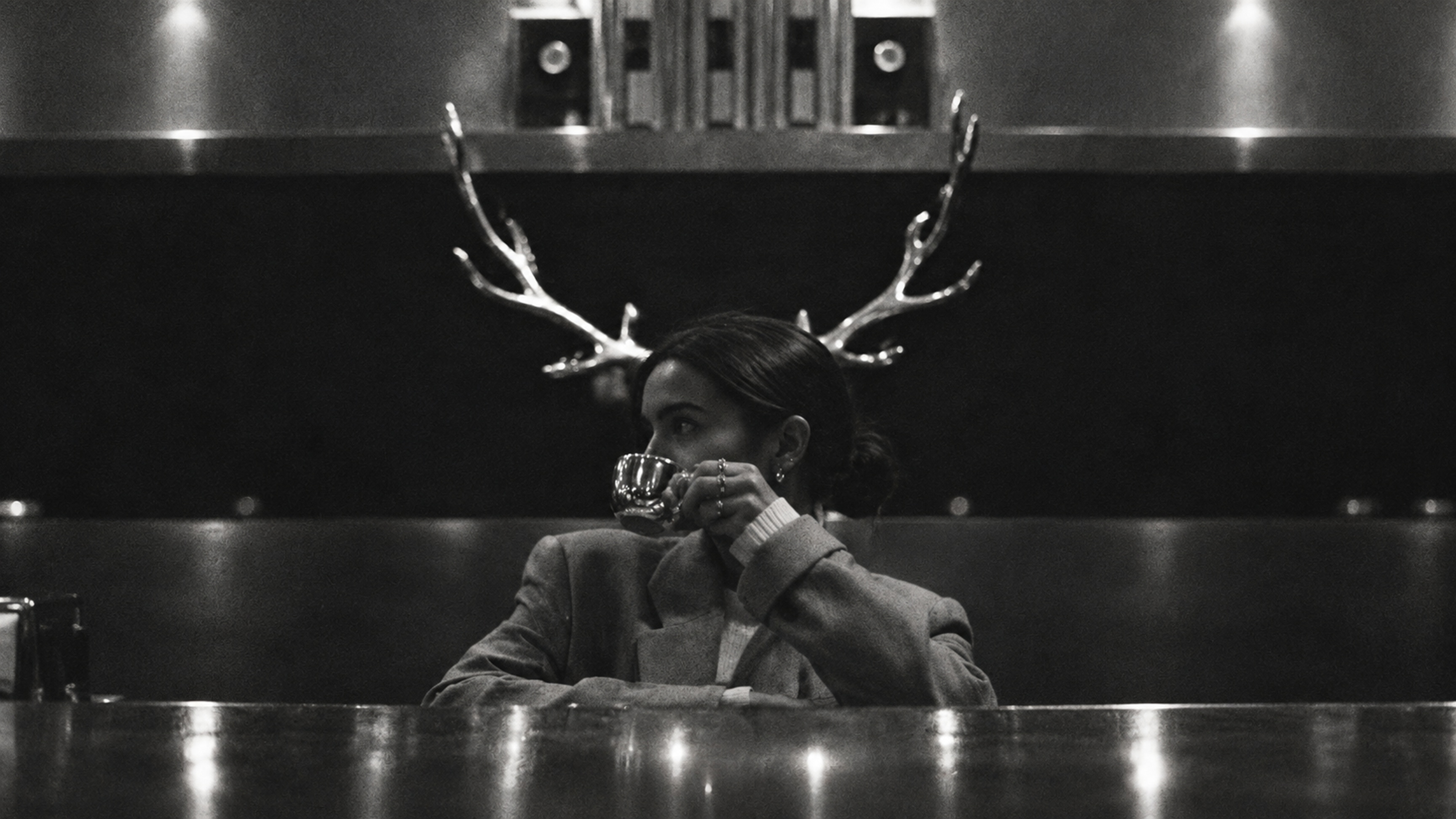 A woman at the bar drinking coffee, metal antlers on the wall behind her head