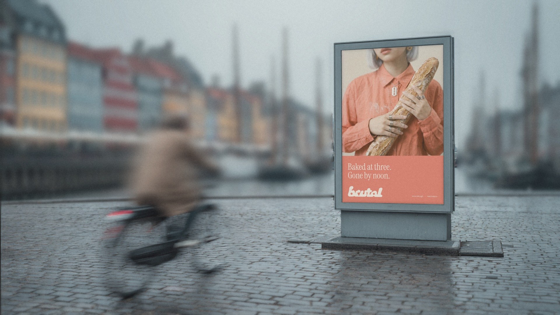 A BRUTAL poster in a lightbox on the quay in Nyhavn, Copenhagen, on a grey morning: Baked at three. Gone by noon. A cyclist passes in a blur.