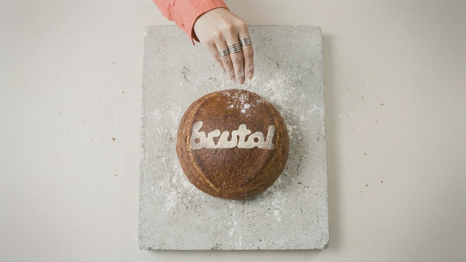 A sourdough loaf on a concrete slab with the BRUTAL logo dusted in flour, a hand sprinkling more from above.